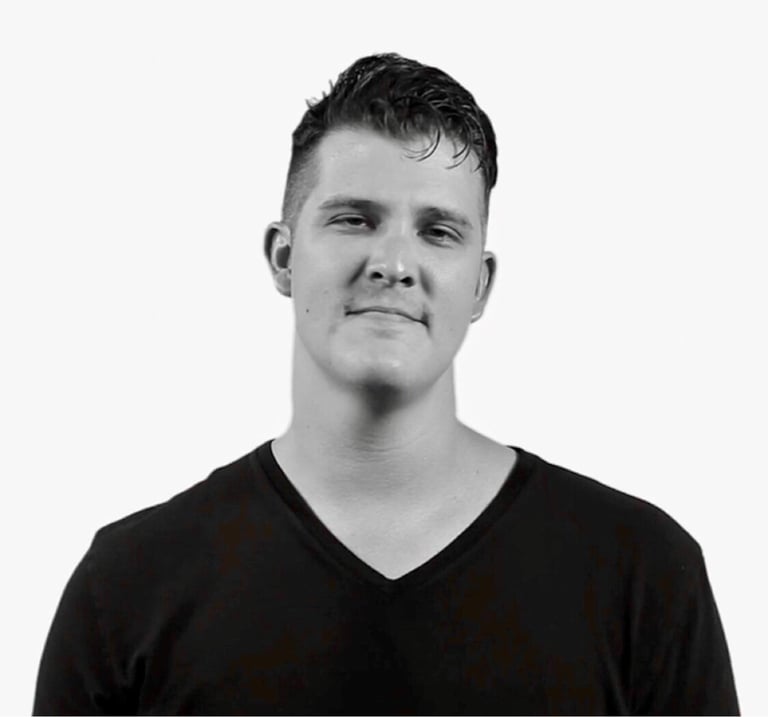 Black and white portrait photograph of a man with short dark hair wearing a black v-neck shirt against a white background